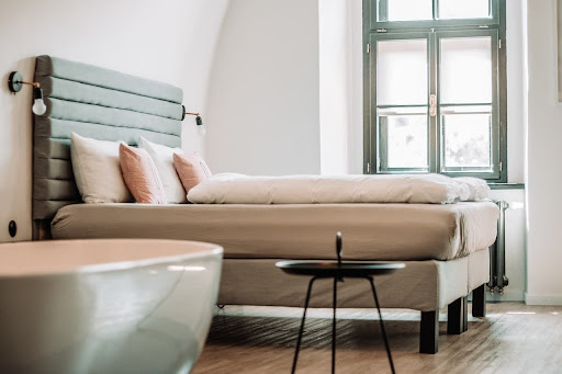 Een elegant ingerichte hotelkamer met een complete luxe boxspring set, een modern gecapitonneerd hoofdbord in pastelgroen, stijlvolle kussens en een vrijstaand bad op de voorgrond.