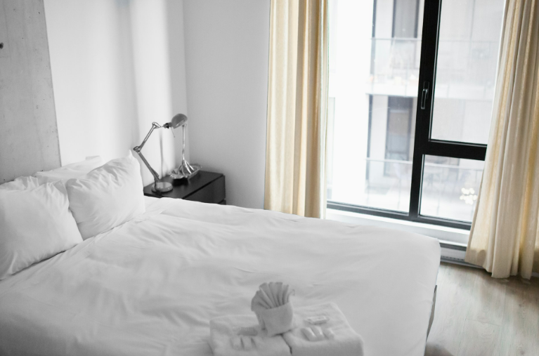 Lichte, moderne hotelkamer met een strak opgemaakt wit bed bij een geopend raam met gordijnen tijdens warm weer.